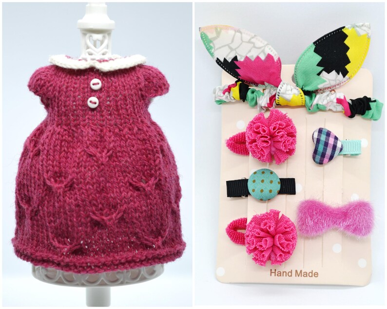 May include: A magenta knitted dress with a white collar and two white buttons. Beside it is a display of hair accessories, including a bow, pom-pom clips, and a patterned headband with bunny ears. The card reads "Hand Made."