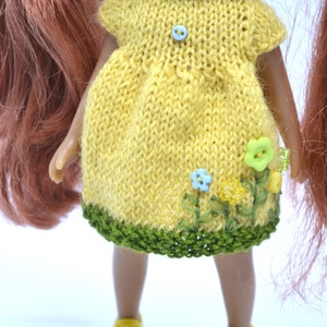 May include: A yellow knitted dress with green trim and embroidered flowers. The dress has a blue button on the front.