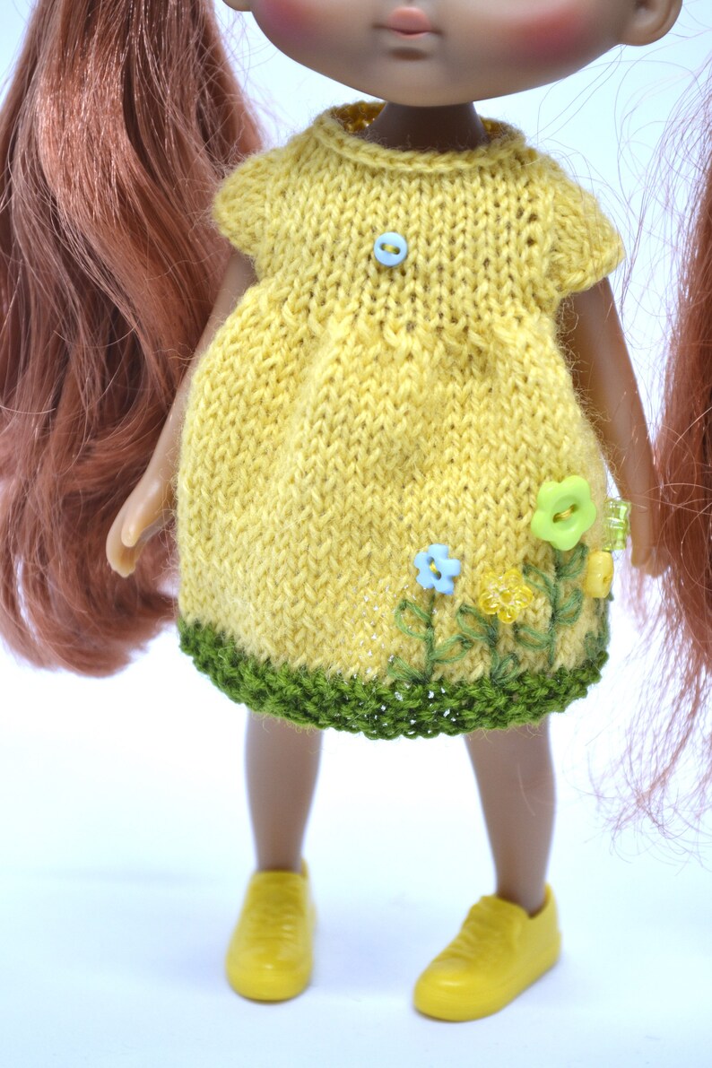 May include: A yellow knitted dress with green trim and embroidered flowers. The dress has a blue button on the front.