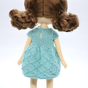 May include: A handmade doll with brown hair in two ponytails. The doll is wearing a light blue knitted dress and brown shoes. The doll is set against a white backdrop.