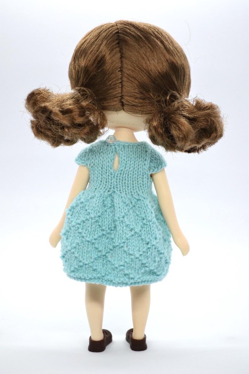 May include: A handmade doll with brown hair in two ponytails. The doll is wearing a light blue knitted dress and brown shoes. The doll is set against a white backdrop.