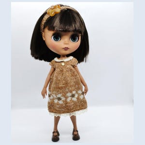 May include: A doll with dark brown hair and large blue eyes, wearing a brown knitted dress with white floral accents and a white lace hem. The doll has a matching headband with a flower and brown shoes.