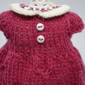 May include: A knitted doll dress in a deep rose colour with a cream-coloured collar. The dress features two small white buttons and is made of a textured yarn. The doll dress is a handmade item.