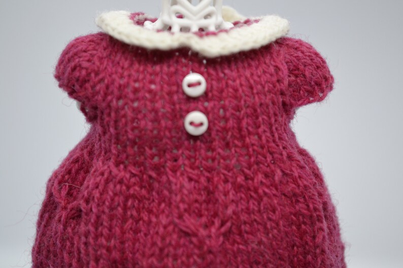 May include: A knitted doll dress in a deep rose colour with a cream-coloured collar. The dress features two small white buttons and is made of a textured yarn. The doll dress is a handmade item.
