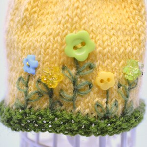 May include: A yellow knitted hat with a green crocheted brim. The hat is decorated with embroidered green leaves and colourful plastic flowers.