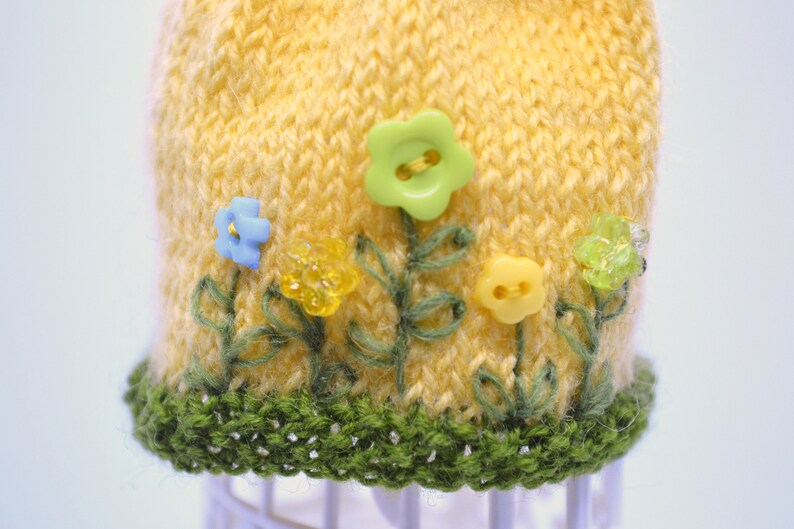 May include: A yellow knitted hat with a green crocheted brim. The hat is decorated with embroidered green leaves and colourful plastic flowers.