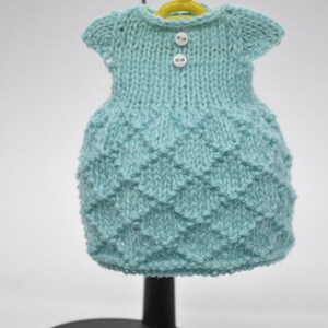May include: A light blue knitted dress with a textured pattern, displayed on a miniature yellow hanger. The dress has short sleeves and two white buttons. The dress is displayed on a black stand.