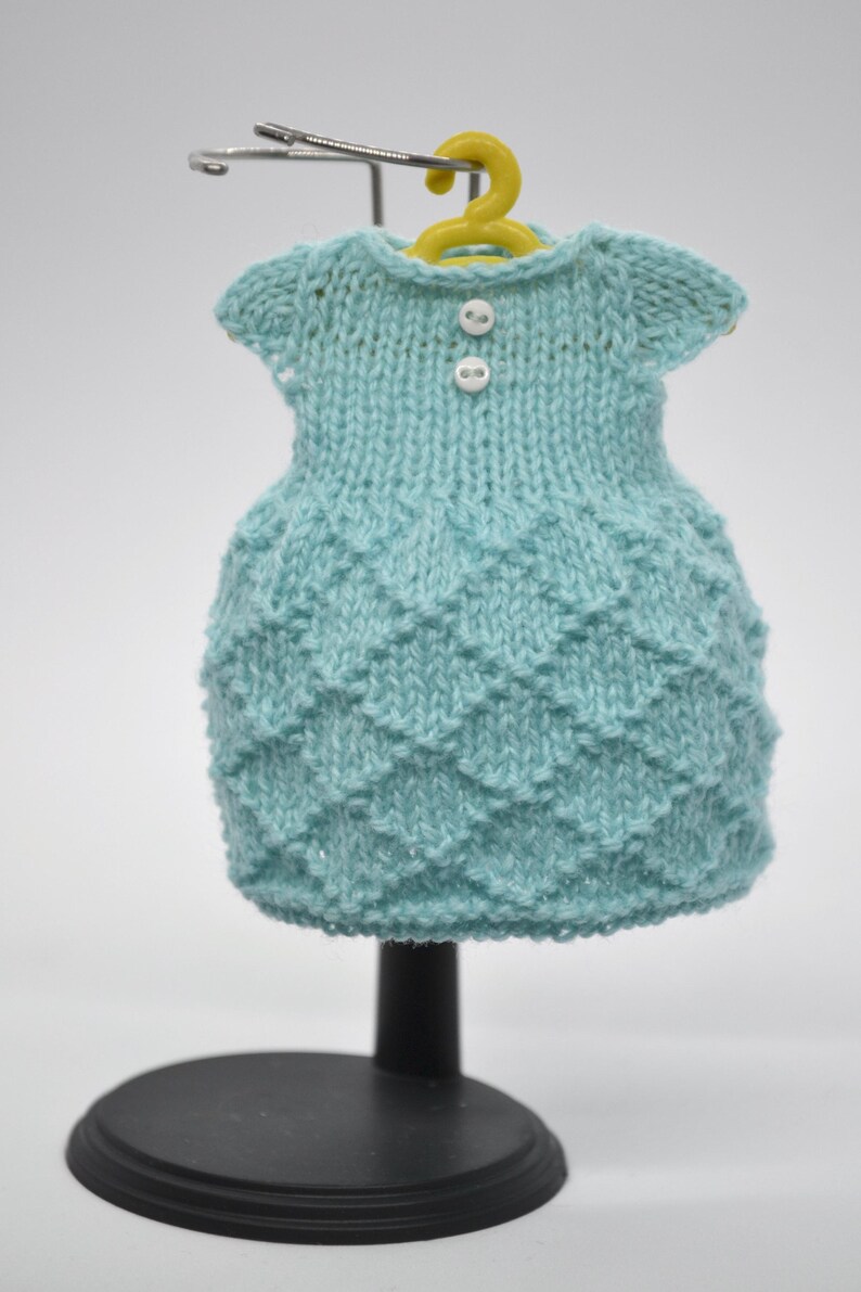 May include: A light blue knitted dress with a textured pattern, displayed on a miniature yellow hanger. The dress has short sleeves and two white buttons. The dress is displayed on a black stand.