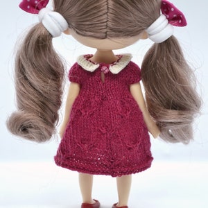 May include: A handmade doll with light brown hair in pigtails, decorated with pink polka dot bows. The doll is wearing a knitted burgundy dress with a white collar and red shoes. The doll is facing away from the camera.