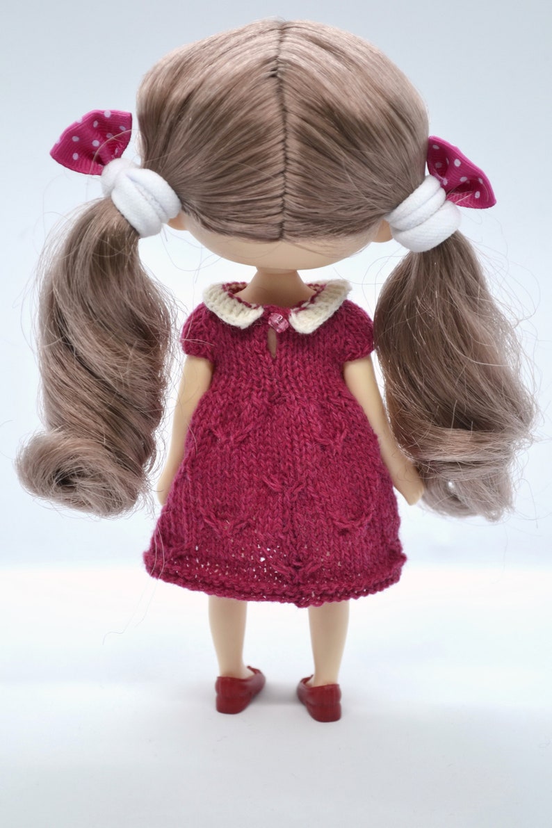 May include: A handmade doll with light brown hair in pigtails, decorated with pink polka dot bows. The doll is wearing a knitted burgundy dress with a white collar and red shoes. The doll is facing away from the camera.