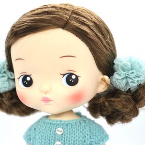 May include: A close-up of a doll with brown hair styled in pigtails, each adorned with a light blue lace flower. The doll has rosy cheeks, a pale pink pout, and is wearing a light blue knitted sweater with white buttons.
