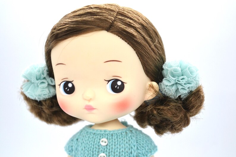 May include: A close-up of a doll with brown hair styled in pigtails, each adorned with a light blue lace flower. The doll has rosy cheeks, a pale pink pout, and is wearing a light blue knitted sweater with white buttons.