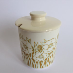 May include: A white ceramic sugar bowl with a lid. The bowl features a floral design with gold and green accents. The flowers are white with a green centre.