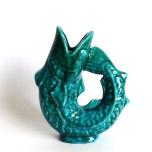 May include: A glossy, deep teal ceramic pitcher shaped like a fish with its mouth open at the top. The body features textured scales and a curved tail that forms a handle-like loop. The glaze has variations in colour intensity across the surface.