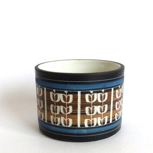 May include: A cylindrical ceramic planter with a white interior and a dark exterior. The outer surface features horizontal bands of black and blue, with a central brown section decorated with a repeating white vertical floral pattern.