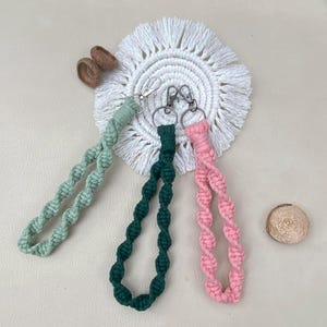 Peut inclure: Trois porte-clés en macramé aux couleurs pastel : vert menthe, vert foncé et rose. Chaque porte-clés a un fermoir argenté et un motif en spirale tissée. Les porte-clés sont présentés sur un dessous de verre blanc à franges.