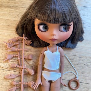 May include: A doll with large eyes and brown hair, wearing a white top and underwear. A set of interchangeable hands is included. The doll has a string attached to a circular object.