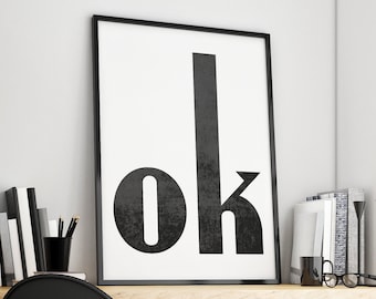 May include: A black and white poster with the word "ok" in bold, block letters. The poster is hanging on a white wall above a wooden desk with a black chair, two potted plants, and a pair of glasses.