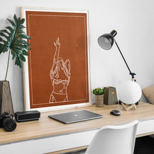 May include: A framed print featuring a minimalist line drawing of a person with their arms raised in the air. The print is on a rust-coloured background and is framed in a natural wood frame. The print is sitting on a desk with a laptop, a lamp, and other office supplies.