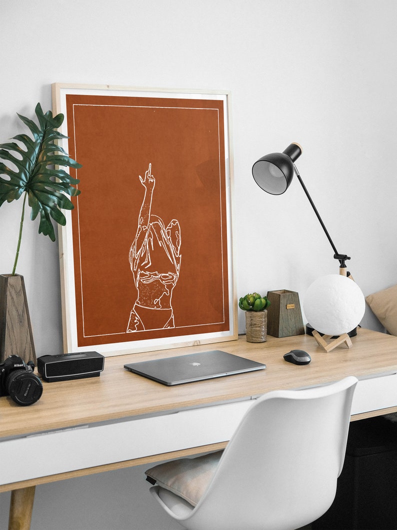 May include: A framed print featuring a minimalist line drawing of a person with their arms raised in the air. The print is on a rust-coloured background and is framed in a natural wood frame. The print is sitting on a desk with a laptop, a lamp, and other office supplies.