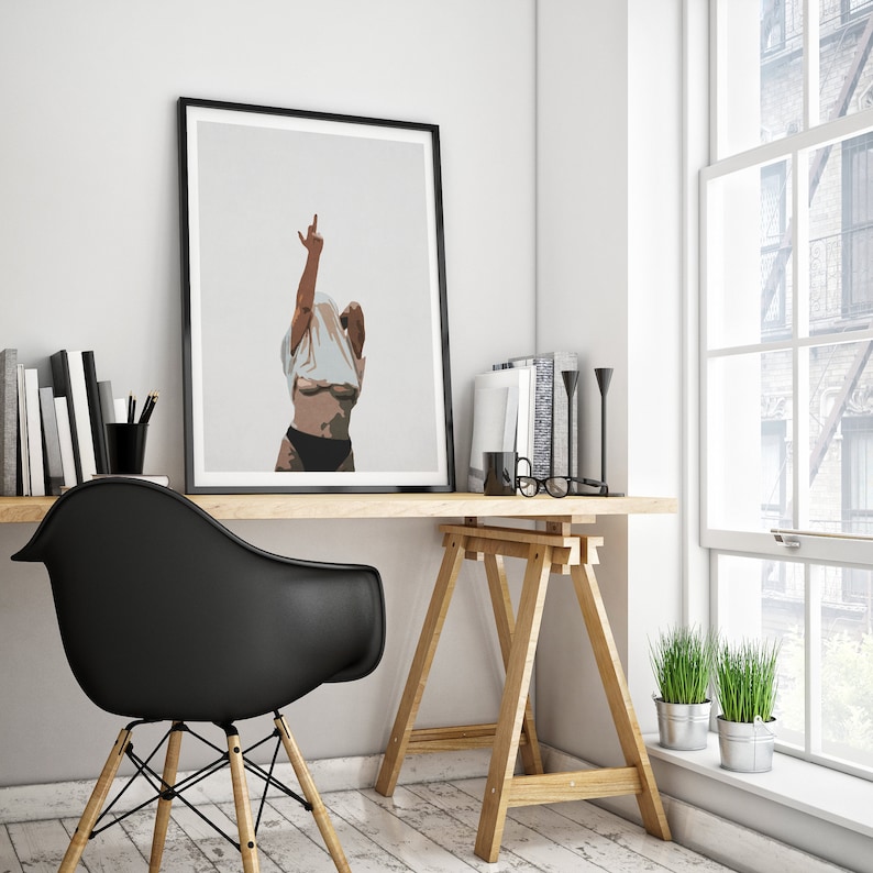 May include: A black framed poster with a white background and a woman in a white shirt with her arms raised. The woman is standing in front of a white wall. The poster is hanging on a wall above a wooden desk.