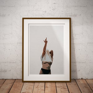 May include: A framed print of a woman in a white t-shirt, with her arms raised and one hand making a peace sign. The background is a light gray.