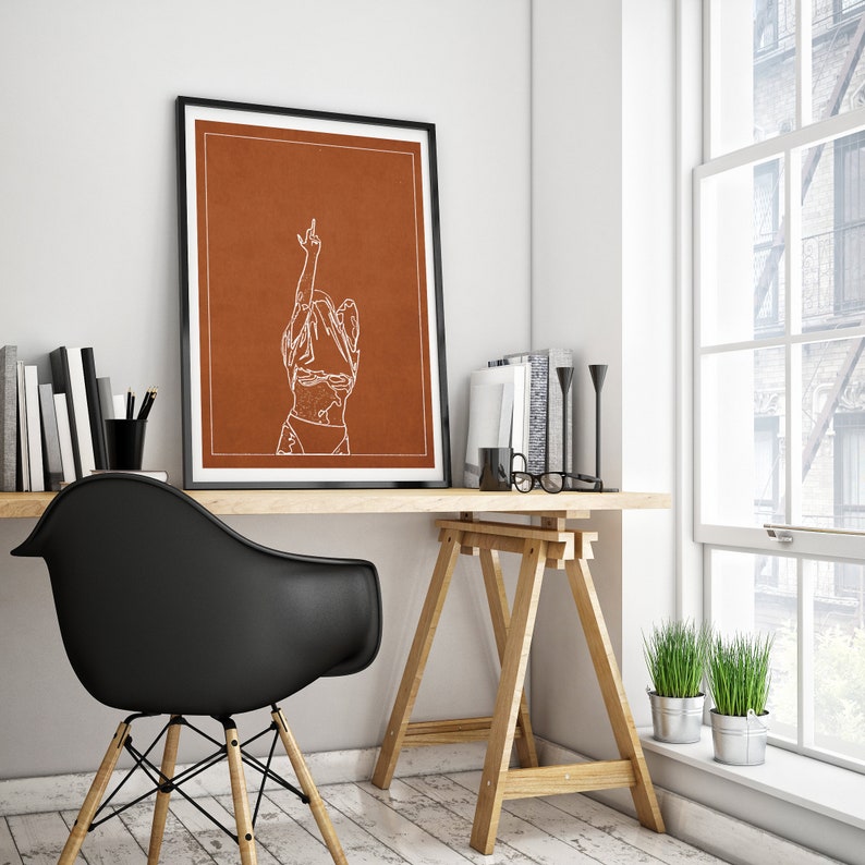 May include: A framed print featuring a minimalist line drawing of a person with their arm raised in the air. The background of the print is a burnt orange colour. The print is hanging on a white wall above a wooden desk.