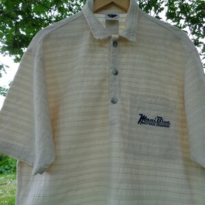 May include: A white and beige striped short-sleeved shirt with a blue embroidered logo that says "Maui Blue Registered Trademark".