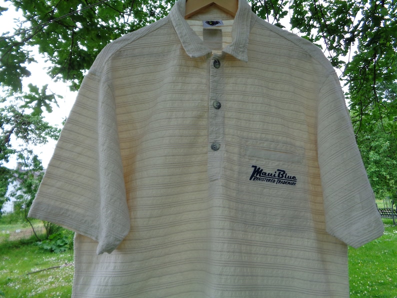 May include: A white and beige striped short-sleeved shirt with a blue embroidered logo that says "Maui Blue Registered Trademark".