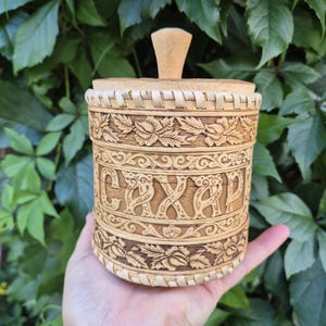 May include: A cylindrical light brown birch bark container with a matching lid and a central wooden knob. The exterior features intricate carved floral patterns and the Cyrillic text CAXAP, which translates to sugar, embossed in a decorative font.