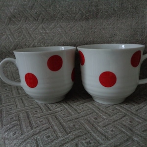 Può includere: Due tazze in ceramica bianca con pois rossi. Le tazze hanno manici e sono appoggiate su una superficie grigia testurizzata.