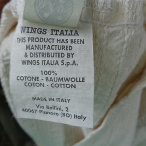 May include: White cotton clothing label with black text that reads "Wings Italia" and "This product has been manufactured & distributed by Wings Italia S.p.A. 100% Cotone - Baumwolle Coton - Cotton Made in Italy Via Bellini, 2 40067 Pianoro (BO) Italy"