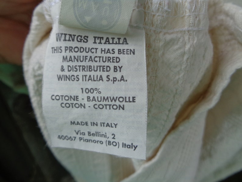 May include: White cotton clothing label with black text that reads "Wings Italia" and "This product has been manufactured & distributed by Wings Italia S.p.A. 100% Cotone - Baumwolle Coton - Cotton Made in Italy Via Bellini, 2 40067 Pianoro (BO) Italy"
