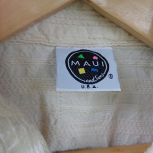 May include: A white cotton shirt with a label that reads "MAUI and Sons" in a black circle with a colorful design. The label also says "U.S.A."
