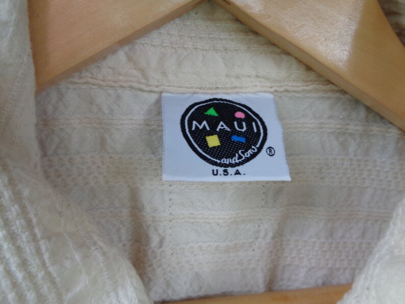 May include: A white cotton shirt with a label that reads "MAUI and Sons" in a black circle with a colorful design. The label also says "U.S.A."