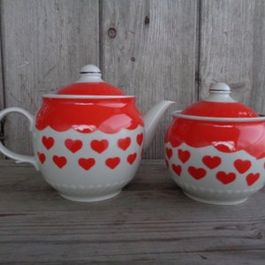 May include: A white teapot and sugar bowl with red lids and red hearts on a white background. The teapot has a handle and a spout. The sugar bowl has a lid.