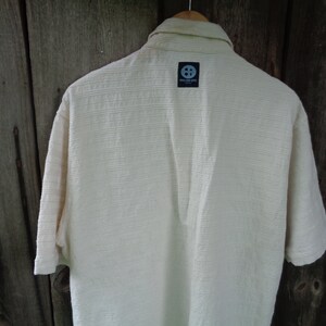 May include: A white short-sleeved button-down shirt with a textured fabric. The shirt has a small black label on the back with the text "The Soul Surf Co."