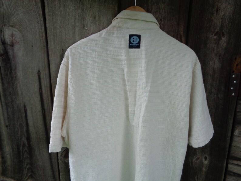 May include: A white short-sleeved button-down shirt with a textured fabric. The shirt has a small black label on the back with the text "The Soul Surf Co."