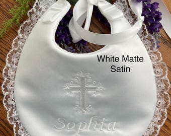 May include: A white matte satin bib featuring a white lace trim along the edge, a white ribbon tie closure, and white embroidery of a cross and the name Sophia. The text White Matte Satin is printed on the surface of the white satin fabric.