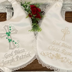 May include: Two white satin baby bibs with lace trim. One bib features a silver cross with green shamrocks and the text "God Bless Joseph Patrick". The other bib features a silver cross and the text "God Bless Claire Rose".