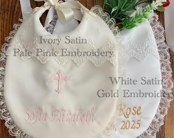 May include: Ivory satin bib with pale pink embroidery, a cross, and the name "Sofia Elizabeth." A white satin bib with gold embroidery features the name "Rose" and the year "2025." Both bibs have lace trim and satin ribbon ties.