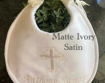 May include: A matte ivory satin baby bib featuring an embroidered cross and the name Anthony James. The bib includes a matching ivory ribbon tie at the top. Text on the image reads Matte Ivory Satin, Anthony James.