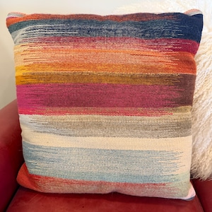 May include: A square decorative pillow with a woven, striped design. The pillow features horizontal bands of color, including blue, orange, pink, and gray. The pillow is placed on a red leather surface.