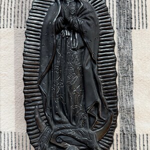 May include: A matte black sculptural wall plaque depicting the Virgin of Guadalupe. The figure stands with hands clasped in prayer, surrounded by a radiant sunburst halo. A small winged angel figure is positioned at the base of the central figure.