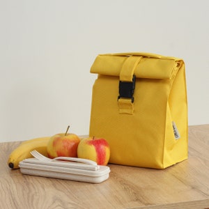 May include: A yellow lunch bag with a black buckle closure. The bag is sitting on a wooden table with two red apples and a banana next to it. The bag has the text "Lunch Bag" printed on the side.