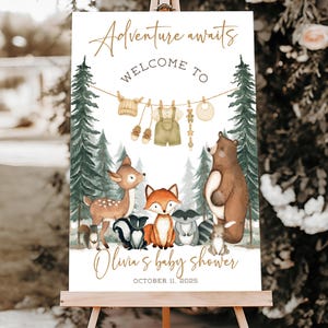May include: A baby shower welcome sign on an easel. The sign features the text "Adventure awaits" and "Welcome to" in gold script. Below are watercolor illustrations of forest animals and baby clothes. The sign reads "Olivia's baby shower" and the date.