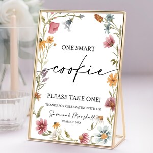 Puede incluir: Un letrero con marco dorado con el texto "ONE SMART cookie PLEASE TAKE ONE!" rodeado por un borde floral. El letrero también incluye el nombre "Savannah Marshall" y "CLASS OF 20XX".