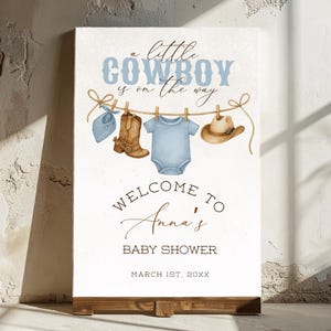 May include: A baby shower sign with the text "a little COWBOY is on the way." The sign features illustrations of cowboy boots, a onesie, a hat, and a bandana hanging on a clothesline. The sign reads "WELCOME TO Anna's BABY SHOWER MARCH 1ST, 20XX."