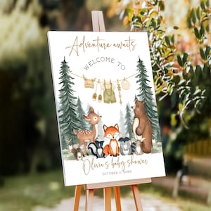 May include: A white sign on an easel with the text "Adventure awaits WELCOME TO" in gold script. Below, a watercolor illustration depicts forest animals and baby clothes. The sign reads "Olivia's baby shower" and the date.