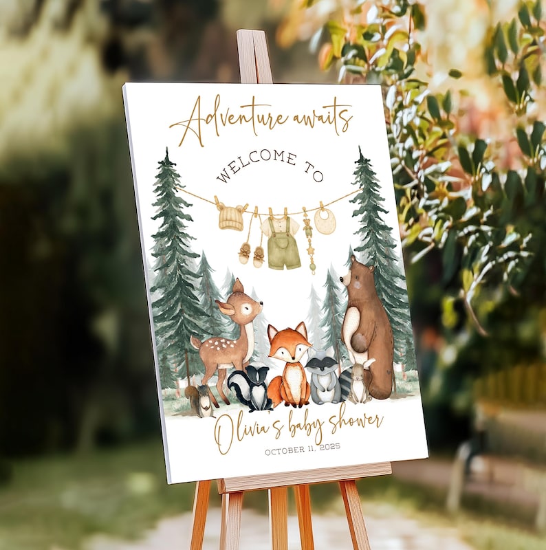 May include: A white sign on an easel with the text "Adventure awaits WELCOME TO" in gold script. Below, a watercolor illustration depicts forest animals and baby clothes. The sign reads "Olivia's baby shower" and the date.